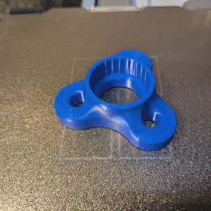 May include: A bright blue, 3D-printed object with a circular center and three rounded arms, each with a hole. The object has a textured interior and sits on a textured surface. The design suggests a tool or component.