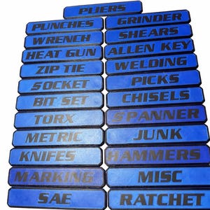 May include: Blue tool labels with white text, including "PLIERS," "WRENCH," "SOCKET," and "KNIFES." Other labels read "GRINDER," "ALLEN KEY," "PICKS," and "HAMMERS." The labels are arranged in rows, ideal for organizing tools.