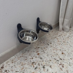 May include: Two wall-mounted pet food bowls. Each bowl is stainless steel and held by a dark blue bracket. The brackets are attached to a white wall. The bowls are empty and ready for food or water. The floor is a speckled terrazzo.