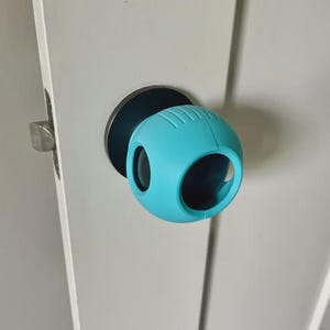 Peut inclure: Un cache-bouton bleu turquoise en forme de sphère avec deux grandes ouvertures. Il est installé sur une poignée de porte avec une base noire. La porte est blanche.