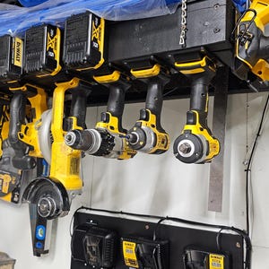 Op de afbeelding: Een verzameling geel en zwart elektrisch gereedschap, waaronder boren, zagen en slijpmachines, is netjes georganiseerd op een zwart rek. Er zijn ook verschillende batterijpakketten zichtbaar. De gereedschappen zijn ontworpen voor bouw- en doe-het-zelfprojecten.