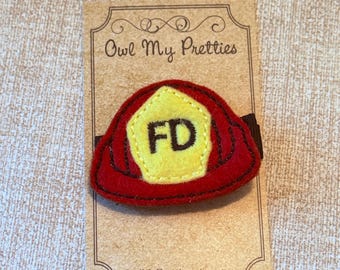 May include: A red felt firefighter helmet hair clip with a yellow center displaying the letters "FD". The hair clip is attached to a brown elastic band and is displayed on a brown card with the text "Owl My Pretties" and "OwlMyPretties.etsy.com".