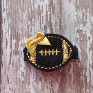 May include: A black felt football with gold stitching and a gold bow. The football is a hair clip.