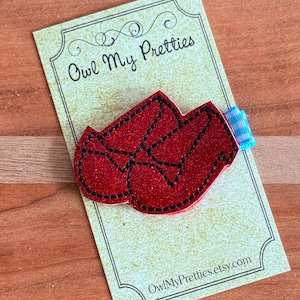 May include: A red glittery hair clip in the shape of ruby slippers, with black stitching details. The clip is on a light green card with the text "Owl My Pretties" and "OwlMyPretties.etsy.com". The background is a wooden surface.