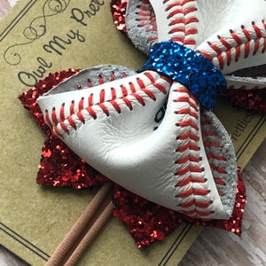 May include: A white leather baseball-themed hair bow with red stitching and a blue glitter center. The bow is attached to a brown elastic headband.