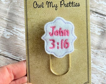 May include: A white and pink felt bookmark with the text "John 3:16" and a gold-colored paper clip. The bookmark is on a brown card with the text "Owl My Pretties" and a website address.