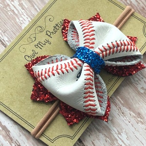 May include: A red and white baseball-themed hair bow with blue glitter accents. The bow is attached to a brown elastic headband.