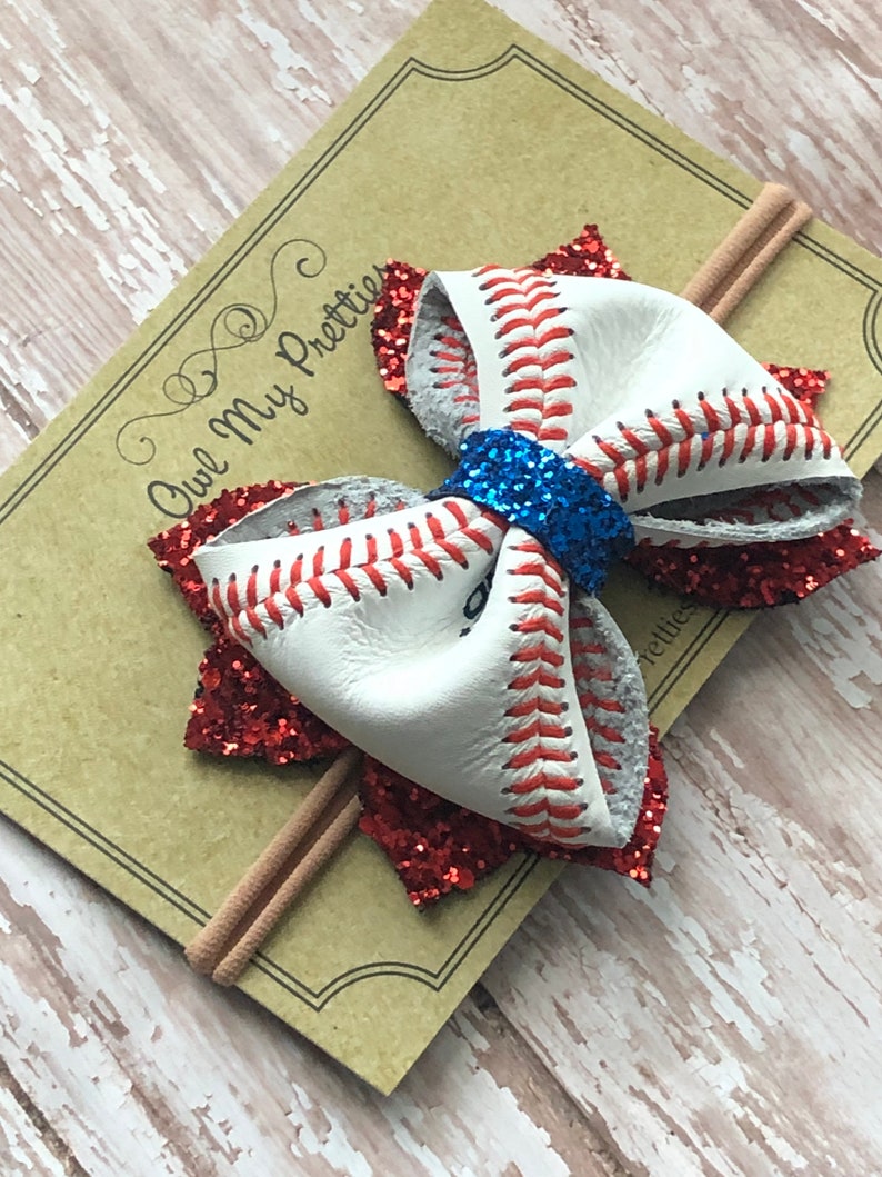 May include: A red and white baseball-themed hair bow with blue glitter accents. The bow is attached to a brown elastic headband.