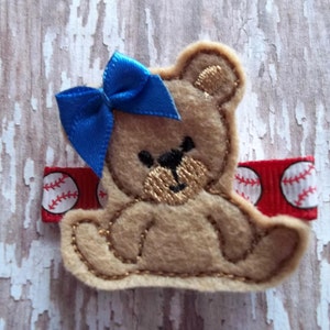 May include: A tan felt teddy bear hair clip with a blue bow. The bear has a stitched face and is attached to a red ribbon with baseballs. A cute accessory for a child.