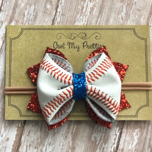 May include: A white and red glitter baseball-themed hair bow with blue glitter in the center, attached to a nude elastic headband. The bow is made of faux leather with red stitching and the text "Owl My Pretties" is visible on the background.