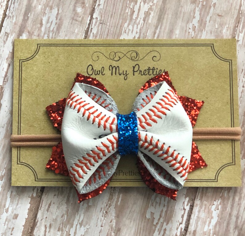 May include: A white and red glitter baseball-themed hair bow with blue glitter in the center, attached to a nude elastic headband. The bow is made of faux leather with red stitching and the text "Owl My Pretties" is visible on the background.