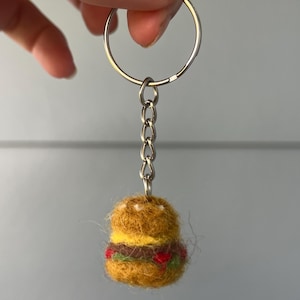 May include: A miniature burger keychain. The burger is made of yellow and brown felt, with layers of yellow, green, and red, resembling cheese, lettuce, and tomato. The keychain has a silver ring and chain.