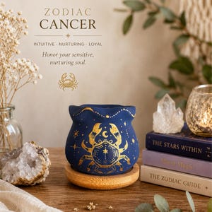 Può includere: Uno scaldacera in ceramica blu scuro con un'illustrazione dorata di un granchio e motivi celesti, poggiato su una base rotonda in legno. Il testo recita: ZODIAC CANCER, INTUITIVE, NURTURING, LOYAL, Honor your sensitive, nurturing soul.