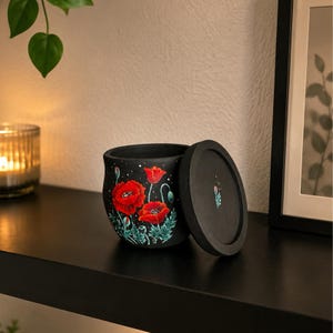 Könnte beinhalten: Ein mattschwarzer zylindrischer Behälter mit passendem Deckel, verziert mit einem handgemalten Motiv aus leuchtend roten Mohnblumen mit grünem Blattwerk und kleinen weißen Punkten. Auf dem Deckel ist eine einzelne kleine Mohnknospe abgebildet.
