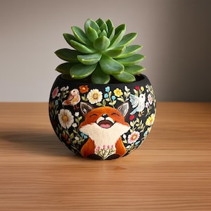 以下が含まれることがあります： 笑顔のキツネのデザイン、カラフルな花、鳥が描かれた黒いセラミックプランター。緑色の多肉植物が入っています。プランターは木製の表面に置かれています。