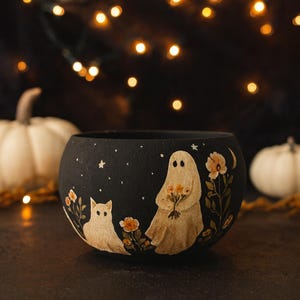 Może przedstawiać: Czarna, okrągła ceramiczna miska z halloweenowym wzorem. Misa przedstawia białego kota, ducha trzymającego kwiaty, gwiazdy i kwiatowe akcenty. Tło jest ciemne, z rozmytymi światłami i białymi dyniami.