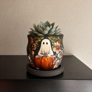 May include: A black ceramic planter featuring a hand-painted white ghost sitting on an orange pumpkin, surrounded by detailed autumn-toned floral illustrations. A green succulent plant is potted inside, and the planter rests on a matching saucer.