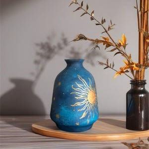 May include: A blue ceramic vase with a gold sun design and small gold stars. The vase is on a wooden tray, with a dark brown glass bottle holding dried plants to the right. The vase is approximately 20 cm tall.