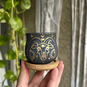 Puede incluir: Una maceta de cerámica negra con un diseño dorado del signo zodiacal Aries. La maceta se apoya en un platillo redondo de madera. El símbolo de Aries está centrado, rodeado de estrellas y lunas crecientes. Diámetro: aprox. 8 cm.