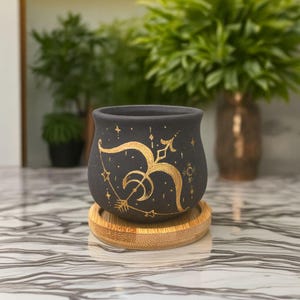 Könnte beinhalten: Ein schwarzer Keramik-Pflanztopf mit einem goldenen Schützen-Design, einschließlich Pfeil und Bogen, Sternen und einem Halbmond. Der Topf steht auf einer runden Bambus-Untertasse. Das Design ist handbemalt.