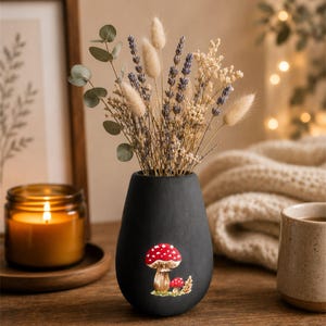 May include: A black ceramic vase with a mushroom design, filled with dried flowers and greenery. The vase has a teardrop shape and sits on a wooden surface. A lit candle and a framed picture are in the background.
