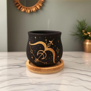 Puede incluir: Maceta de cerámica negra con un diseño dorado de Sagitario, que incluye un arco y flecha, estrellas y sol. La maceta se apoya en un posavasos redondo de bambú. Un espejo dorado en el fondo.