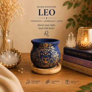 May include: A dark blue ceramic pot featuring a gold hand-painted lion design, resting on a circular wooden base. The pot is surrounded by books titled The Stars Within, Moon Magic, and The Zodiac Guide, along with crystals and a candle.