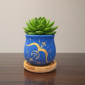 May include: A small, blue ceramic planter with a gold Sagittarius design, holding a green succulent plant. The planter sits on a round, wooden coaster. The design includes a bow and arrow, stars, and a crescent moon.