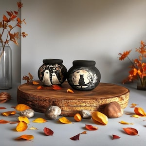 May include: Two black, round ceramic pots with Halloween-themed designs. One pot features two white ghosts under a starry sky, the other a black cat on a branch in front of a moon. The pots sit on a wooden slab with autumn leaves.