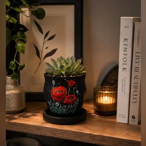 May include: A black ceramic planter featuring a hand-painted design of vibrant red poppy flowers and green foliage. The round pot sits on a matching black saucer and contains a green succulent plant with thick, pointed leaves arranged in a rosette.