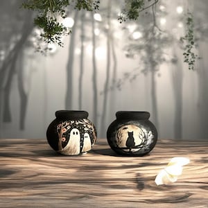 Puede incluir: Dos macetas negras de cerámica con diseños pintados a mano con temática de Halloween. Una maceta presenta dos fantasmas blancos bajo un árbol, la otra un gato negro en una rama. Las macetas están sobre una superficie de madera.