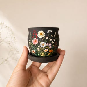 Puede incluir: Una pequeña maceta de cerámica negra mate con su plato a juego, que presenta un diseño botánico pintado a mano de varias flores silvestres en tonos blanco, rosa, amarillo y naranja, con tallos y hojas verdes alrededor del exterior.