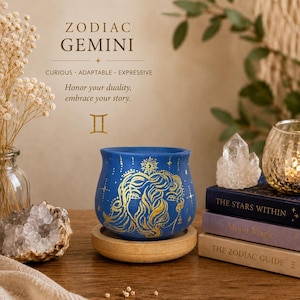 May include: A blue ceramic pot featuring a gold-colored Gemini zodiac illustration, resting on a circular wooden base. The text reads ZODIAC GEMINI, CURIOUS ADAPTABLE EXPRESSIVE, Honor your duality, embrace your story, and the Gemini symbol.
