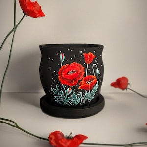 May include: A matte black cylindrical plant pot with a matching saucer, featuring a hand-painted design of vibrant red poppy flowers, green foliage, and small white dots scattered across the surface to resemble a starry night sky.