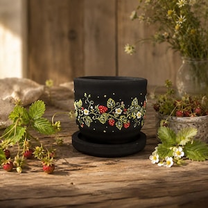 Puede incluir: Una maceta de cerámica negra mate con su plato a juego, decorada con un diseño pintado a mano de hojas de fresa verdes, flores blancas y fresas rojas que rodean el exterior, acentuado con pequeñas estrellas amarillas.