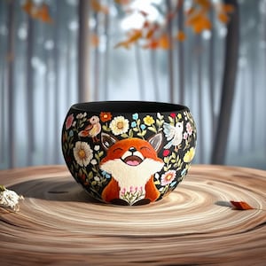 May include: A black ceramic bowl with a hand-painted design featuring a smiling fox, birds, and colourful flowers. The bowl is set on a wooden surface, showcasing the intricate details of the artwork. The overall theme is nature and woodland creatures.