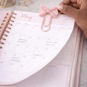 May include: A pink planner with a rose gold spiral binding, open to the May calendar. A decorative pink bow paper clip is attached to the page. The calendar has handwritten notes and dates.
