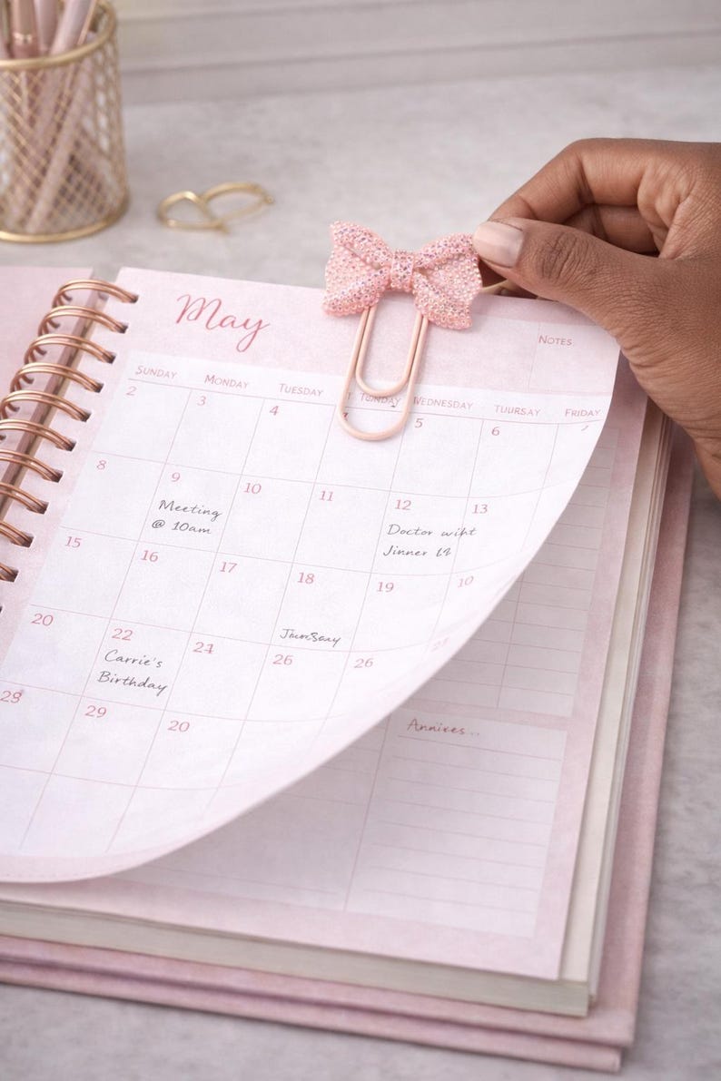 May include: A pink planner with a rose gold spiral binding, open to the May calendar. A decorative pink bow paper clip is attached to the page. The calendar has handwritten notes and dates.