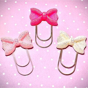 May include: Three decorative bow-shaped paper clips. One is bright pink, one is light pink, and one is off-white. Each bow is embellished with glitter and attached to a gold-coloured metal paper clip. The background is pink with white polka dots.
