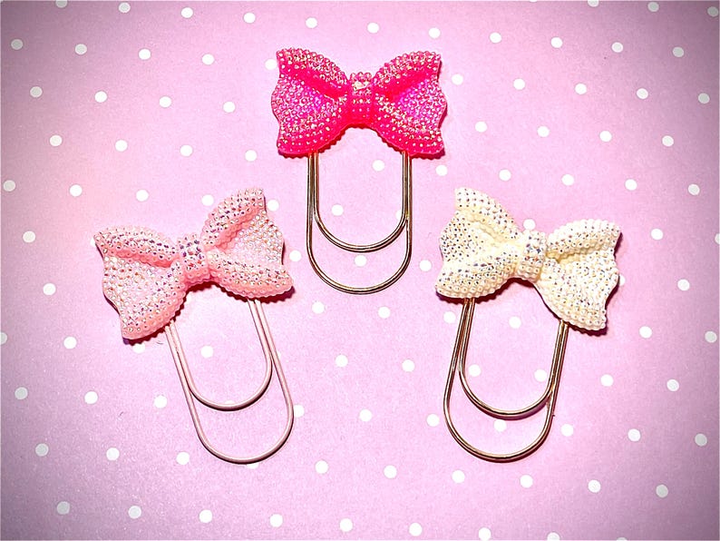 May include: Three decorative bow-shaped paper clips. One is bright pink, one is light pink, and one is off-white. Each bow is embellished with glitter and attached to a gold-coloured metal paper clip. The background is pink with white polka dots.