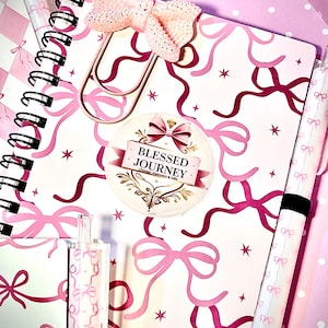 May include: A pink and white notebook with a bow and ribbon design, a decorative bow paperclip, and a sticker reading "Blessed Journey Christian Stationery & Gifts." Also visible are matching pens and a highlighter.