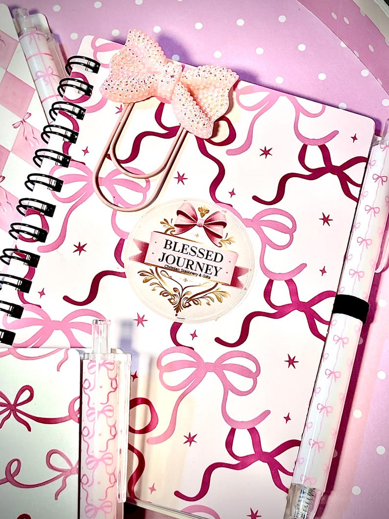 May include: A pink and white notebook with a bow and ribbon design, a decorative bow paperclip, and a sticker reading "Blessed Journey Christian Stationery & Gifts." Also visible are matching pens and a highlighter.