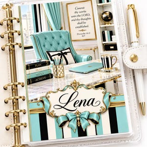 May include: A white ring-bound planner with gold hardware and a white pen, featuring a cover image of a turquoise office with a desk, a chair, a stack of books labeled Plan, Pray, Focus, Achieve, and a sign reading Commit thy works unto the LORD.