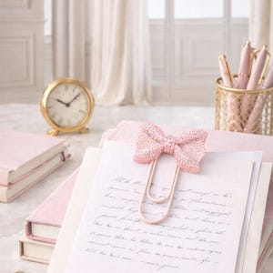 May include: A pink glitter bow paper clip securing papers with handwritten script. A gold clock, pink pens in a wire basket, and pink notebooks are also visible.
