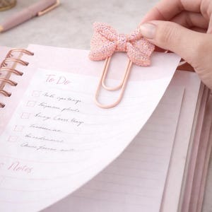 May include: A pink glitter bow paper clip is attached to a pink notebook with a spiral binding. The notebook is open to a page with a handwritten "To Do" list. A pink pen and a gold clock are in the background.