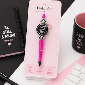 May include: A pink ballpoint pen with black and white cow-print beads and a round black charm featuring the text Faith Over Fear. The pen is mounted on a light pink display card labeled Faith Pen with icons and text including Made With Love.