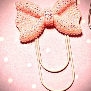 May include: A pink bow-shaped paper clip adorned with iridescent rhinestones. The bow is attached to a light pink paper clip. The background is a pink surface with white polka dots. This decorative paper clip is ideal for stationery and office supplies.