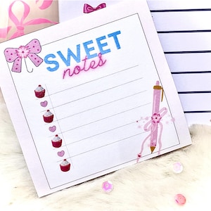 May include: A white notepad with the words "SWEET notes" in blue and pink. The notepad is decorated with pink bows, cupcakes, and hearts. A pink pen is also featured on the notepad.