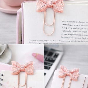 May include: Glittery pink bow-shaped paper clips. The clips are attached to pages of a notebook and a book. The words "Must Do" are written on one of the pages. A pink cup and saucer are in the background.