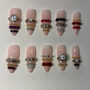 May include: A set of ten artificial nails with a light pink base. Each nail is adorned with various rings, including silver, gold, and beaded designs. Some nails feature a small watch face. The rings are in various colors, including red, black, and green.